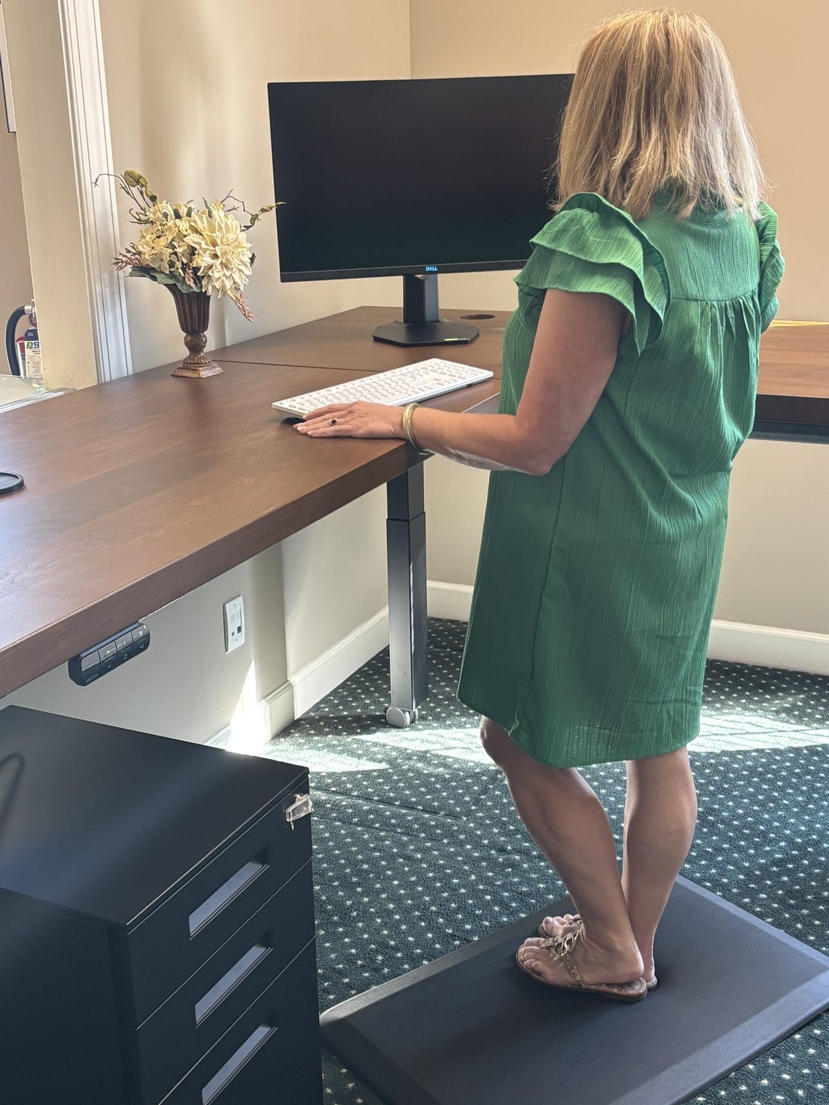 A staff member standing at the desk in daily use, hand on the keyboard, with the monitor at eye height and the desk at standing height on the anti-fatigue mat.