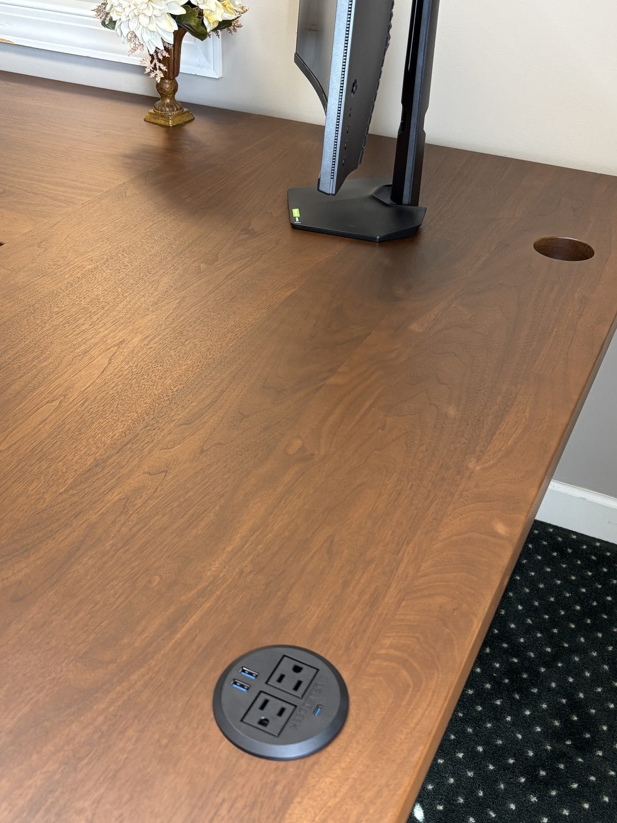 Close-up of the solid walnut desktop grain with a round power grommet holding two outlets and two USB ports built into the surface, next to a monitor stand.