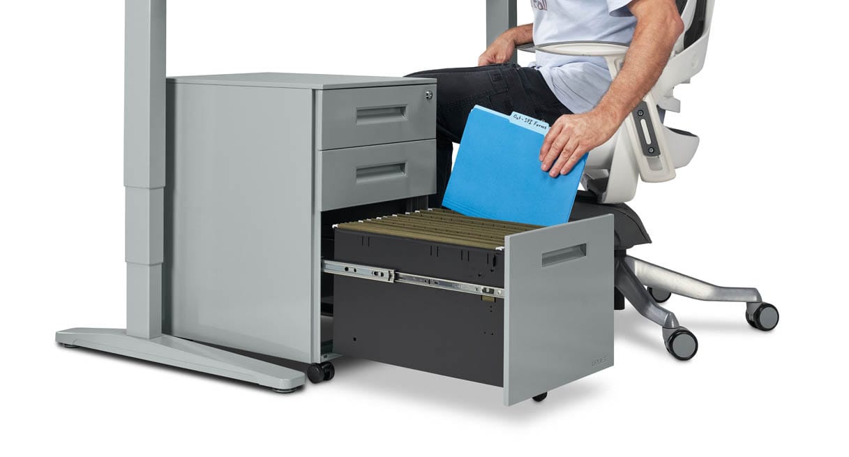 A gray three-drawer file cabinet beside the leg of a gray standing desk with its large bottom drawer pulled out on metal glides, holding green hanging folders while a seated person slides a blue file folder in.