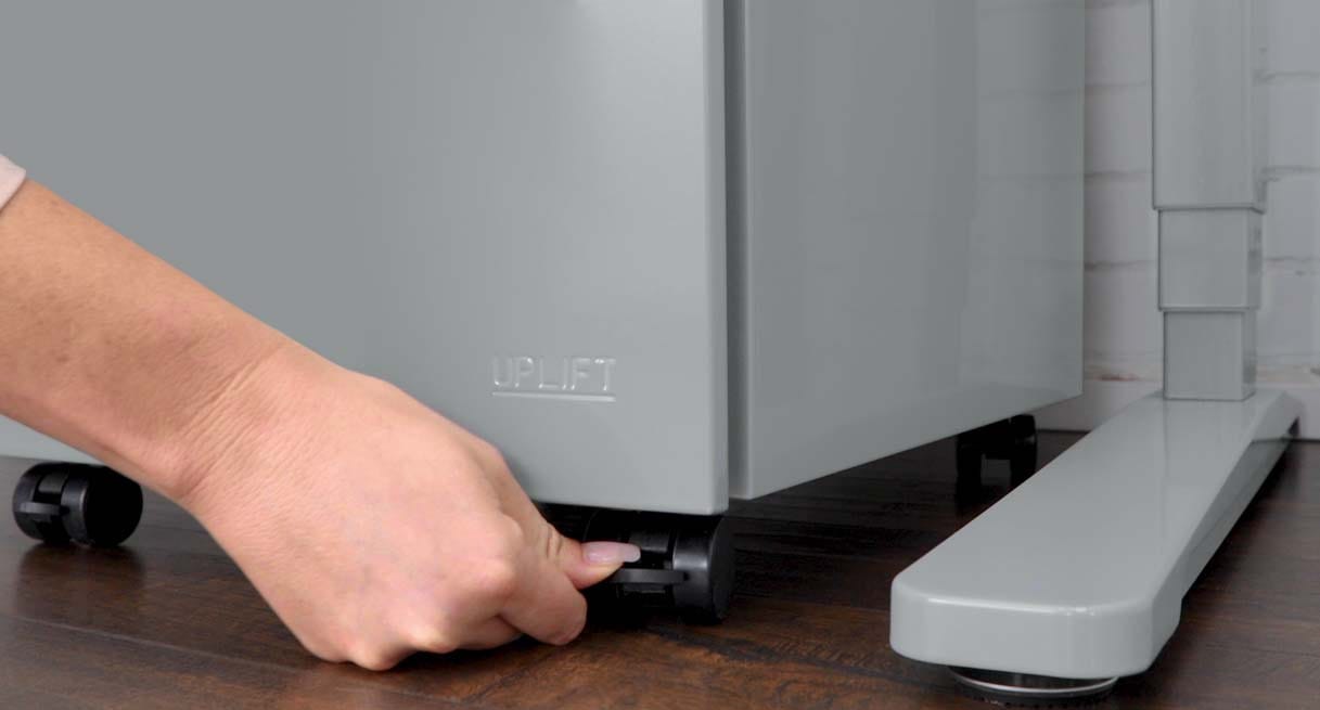 A close-up of a hand pressing the lock tab on a black caster at the front corner of a gray file cabinet embossed with the UPLIFT name, beside the foot of a gray desk frame on a wood floor.