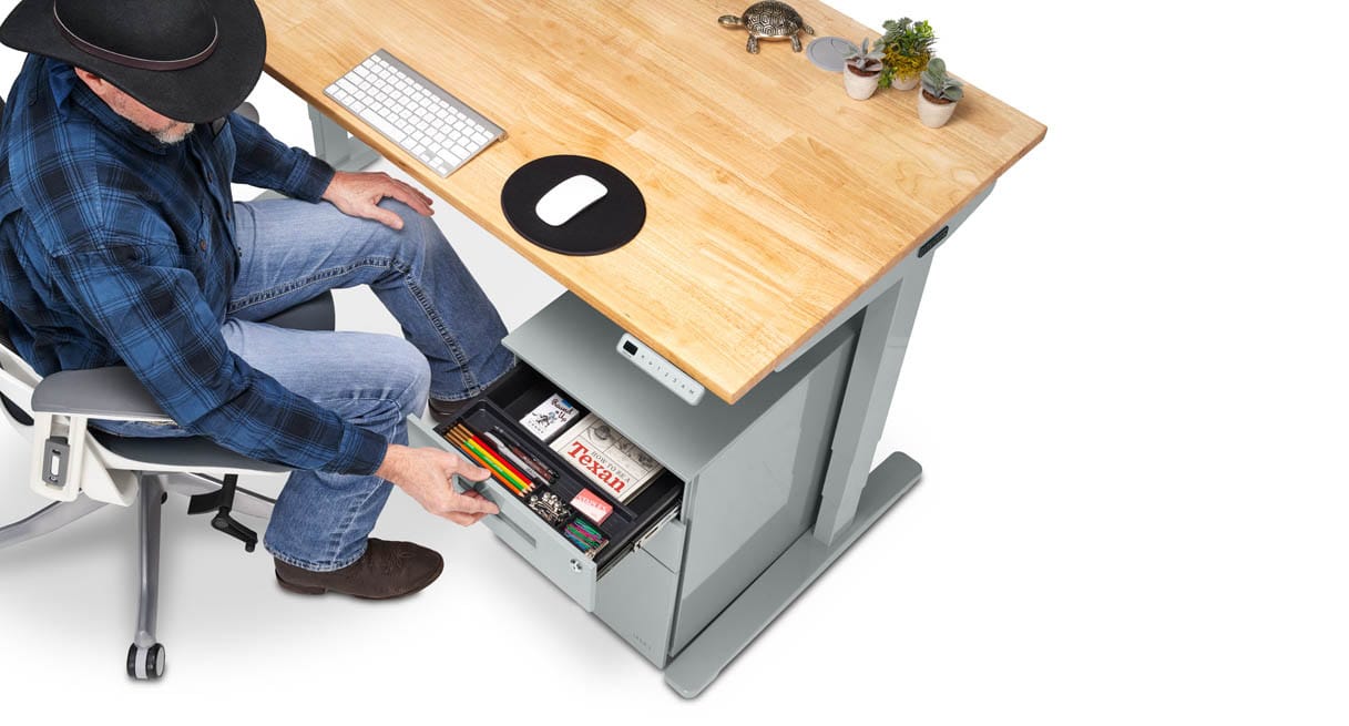 A seated person in a plaid shirt opens the top drawer of a gray file cabinet under a light wood standing desk, showing a black tray filled with pencils, pens and small office items.