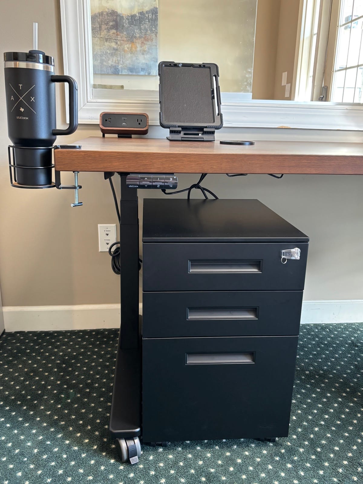Black three-drawer UPLIFT file cabinet standing under a walnut reception desk, with a key and clear key tag in the top-drawer lock, a desk control keypad above it, a tablet stand and power strip on the desktop, and a tumbler in a clamp-on cup holder