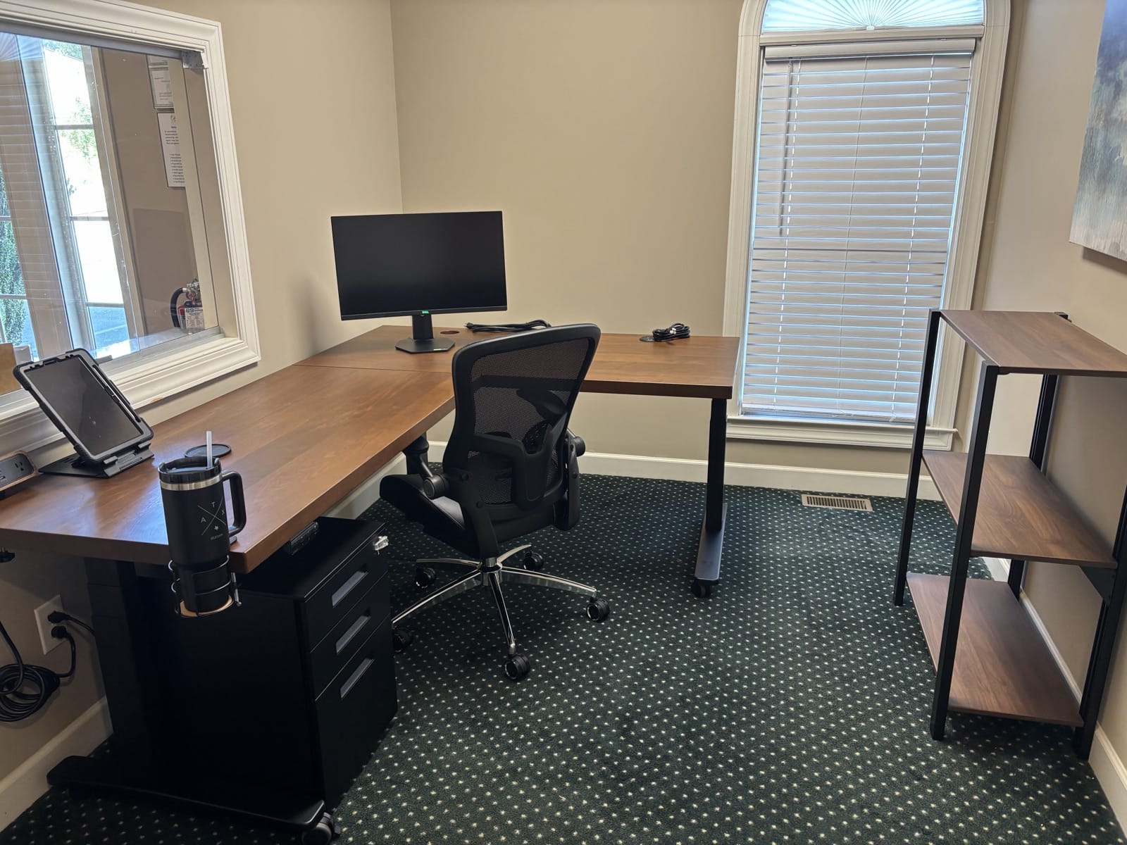 The reception room with the walnut L-shape desk on a black frame at left, a black file cabinet under the desk, a mesh office chair, and the black-framed Sequence Shelving unit with walnut shelves against the wall at right.